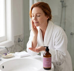 Woman with red hair applying Pūraka Cleansing Wash at bathroom basin in morning light