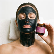 Woman lying down with Pūraka Activated Charcoal Deep Cleansing Mask applied to face holding amber glass jar