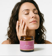 Woman with radiant glowing skin touching her face beside Pūraka Active Day Cream jar