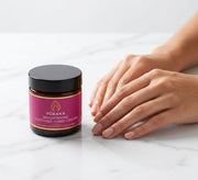 Pūraka Brightening Soothing Hand Cream in amber glass jar on white marble surface beside soft luminous hands
